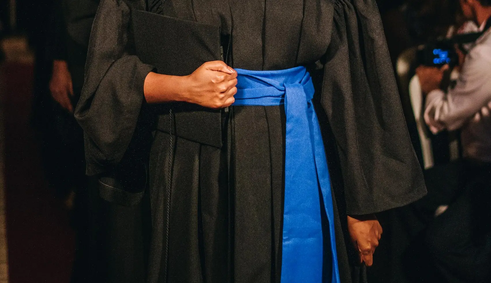 Foto em close-up de uma mulher negra vestida de toga preta de formatura, com faixa azul enrolada na cintura. Ela segura uma pasta preta junta ao corpo. Atrás dela, há uma fila de mais pessoas formadas e, ao lado, há um fotógrafo.