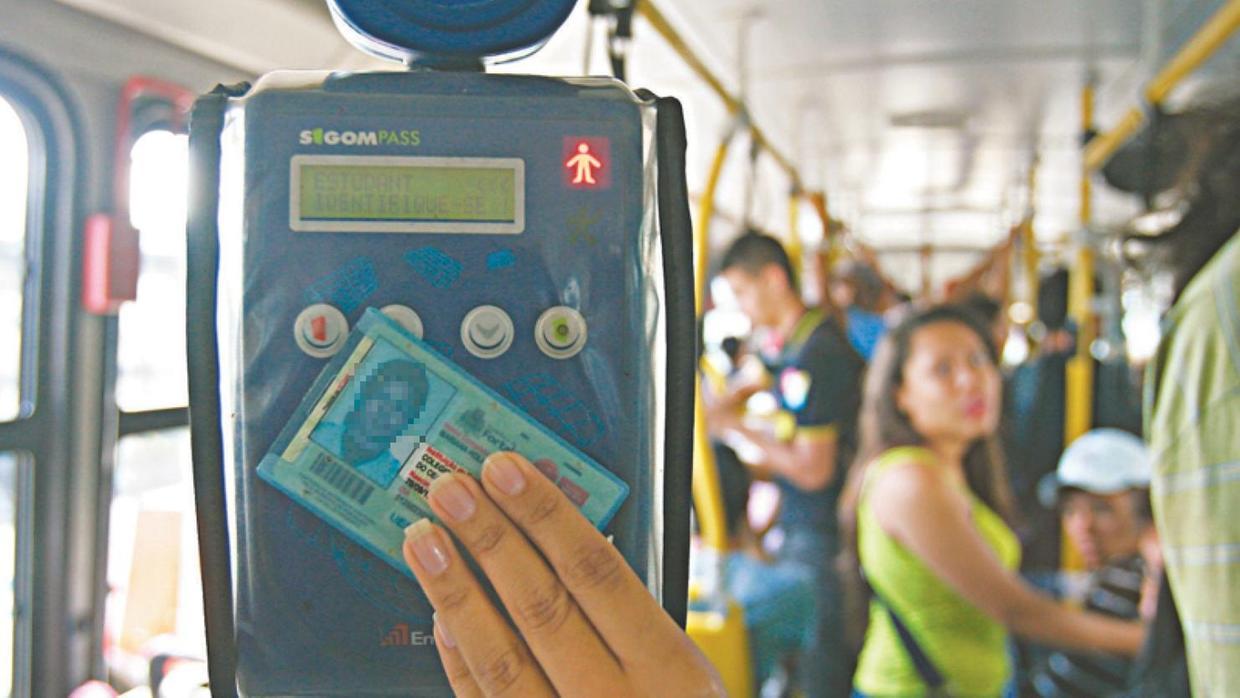 Mão de uma pessoa segura uma carteira de estudante azul e aproxima o documento do validador eletrônico dentro de um ônibus lotado, onde passageiros aparecem desfocados ao fundo.
