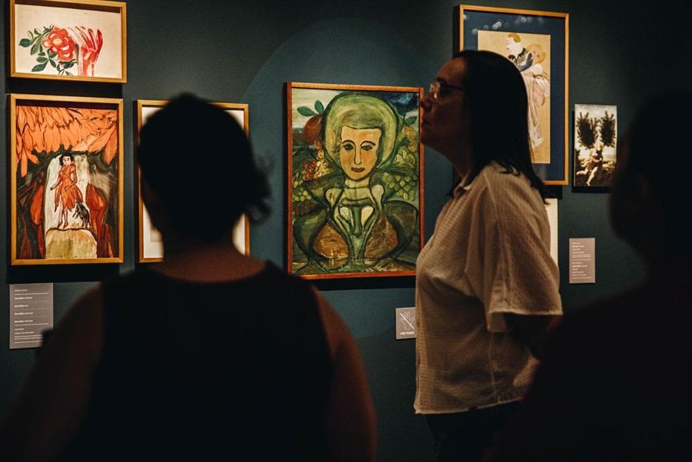 Na imagem, três pessoas estão em uma exposição de arte olhando para pinturas emolduradas penduradas em uma parede azul-escura. A pintura central é um retrato estilizado e colorido de uma figura de pele clara e cabelo verde, emoldurado em madeira. As duas pessoas em primeiro plano estão em silhueta, e a terceira pessoa à direita, de óculos e camisa branca, está mais iluminada, olhando para cima, para a pintura. O título da exposição visível na legenda da imagem é 