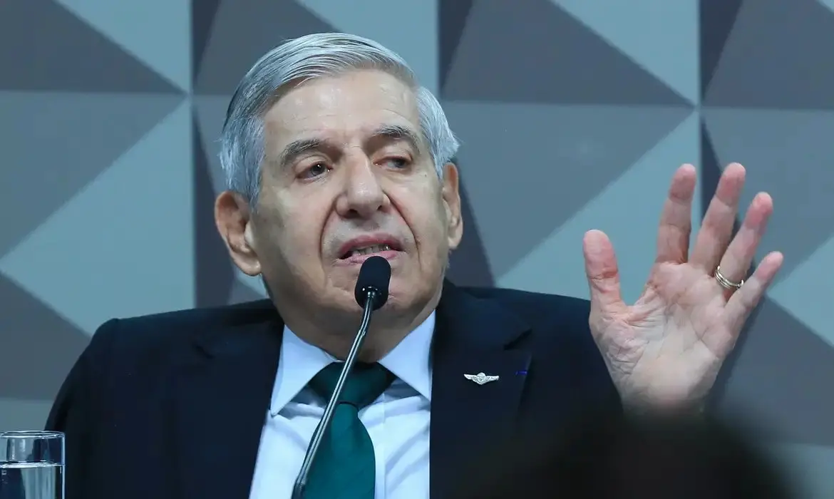 General Heleno de terno e gravata verde, com cabelos grisalhos, falando em um microfone e gesticulando com a mão esquerda levantada, em um ambiente formal.