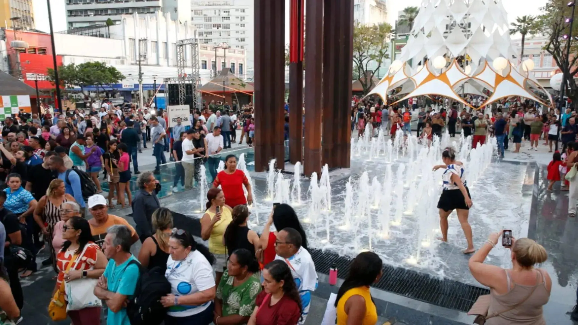 Praça do Ferreira no Centro de Fortaleza. Imagem mostra a fonta interativa, com pessoas se molhando e outras ao redor olhando e fotografando.