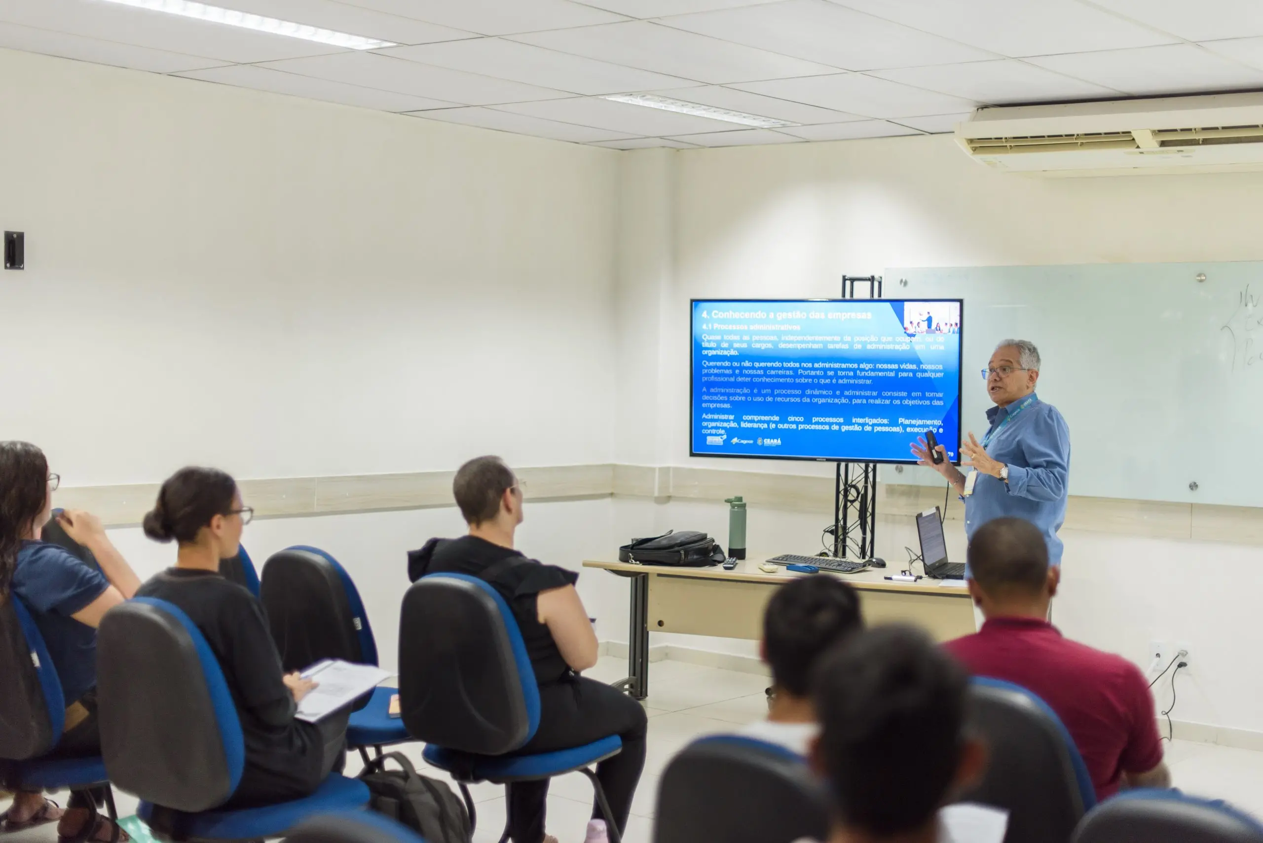 Na imagem, uma sala com uma pessoa falando enquanto olha para um slide, assistindo estão pessoas sentadas em cadeiras.