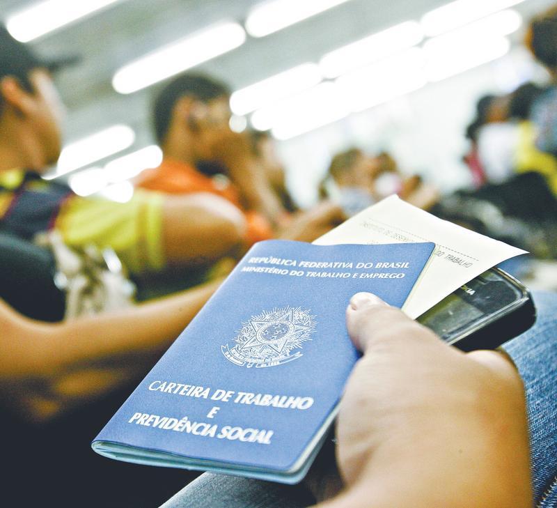 Mão de pessoa segurando a Carteira de Trabalho e Previdência Social (CTPS) brasileira em um ambiente com outras pessoas ao fundo.