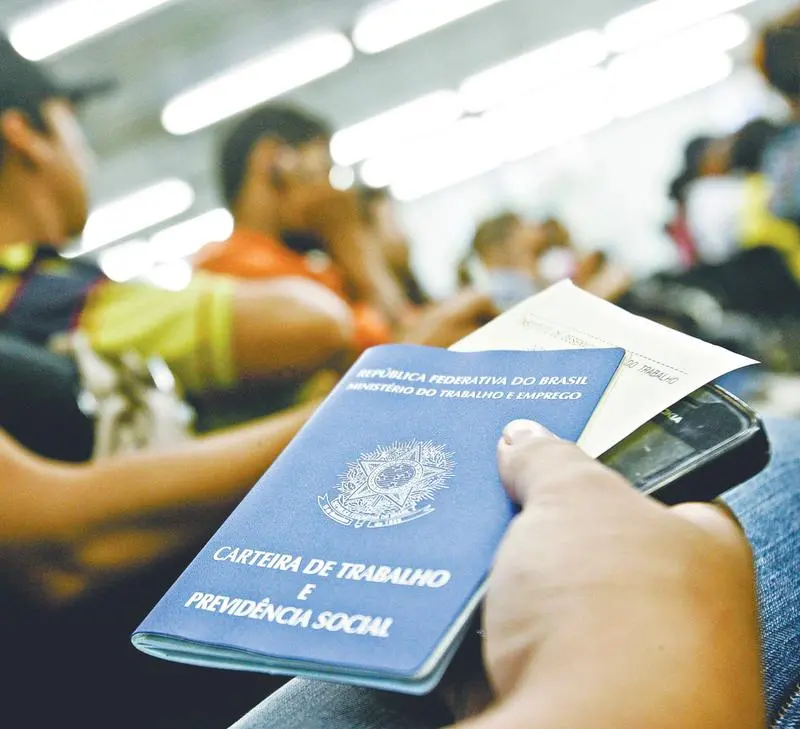 Mão de pessoa segurando a Carteira de Trabalho e Previdência Social (CTPS) brasileira em um ambiente com outras pessoas ao fundo.