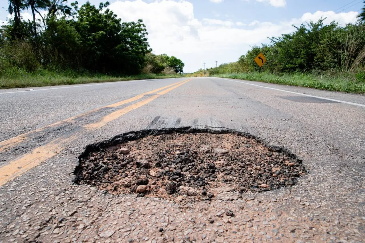 Buraco raso e grande aberto no meio de uma rodovia vazia com grama e árvores verdes nas laterais.