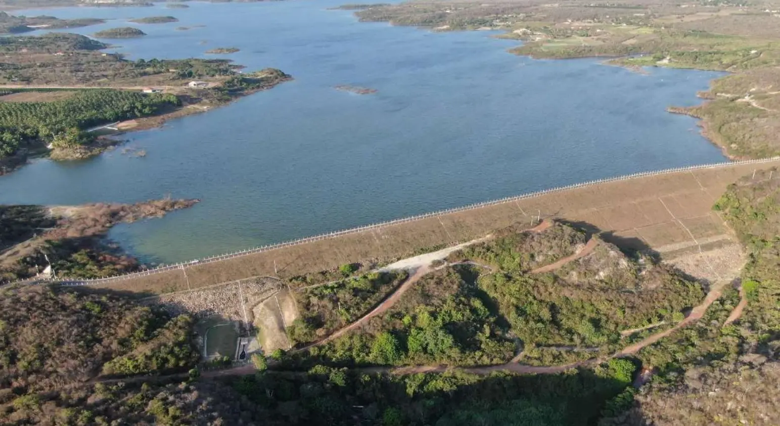 Vista aérea de uma vasta represa com uma barragem de terra, cercada por vegetação esparsa e paisagem semiárida.