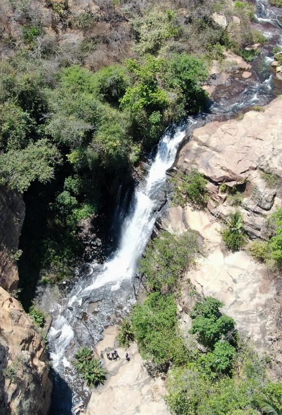 A foto mostra uma queda-d’água alta em meio a um vale estreito em Ubajara, no Ceará, cercado por vegetação nativa típica de clima semiárido de altitude. O rio desce por uma fenda rochosa, formando um poço na parte inferior, onde é possível ver um pequeno grupo de pessoas reunidas sobre uma clareira de pedra. As encostas ao redor têm áreas mais secas misturadas com manchas verdes mais densas.