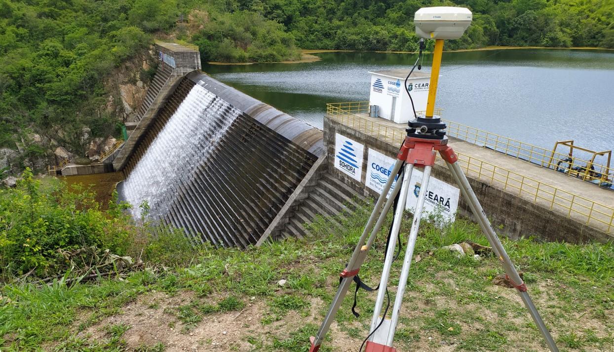 Vertedouro do açude Germinal em operação, com água descendo por uma estrutura em degraus. Em primeiro plano, há um equipamento geodésico sobre tripé, usado para monitoramento. O reservatório fica ao fundo, cercado por vegetação.