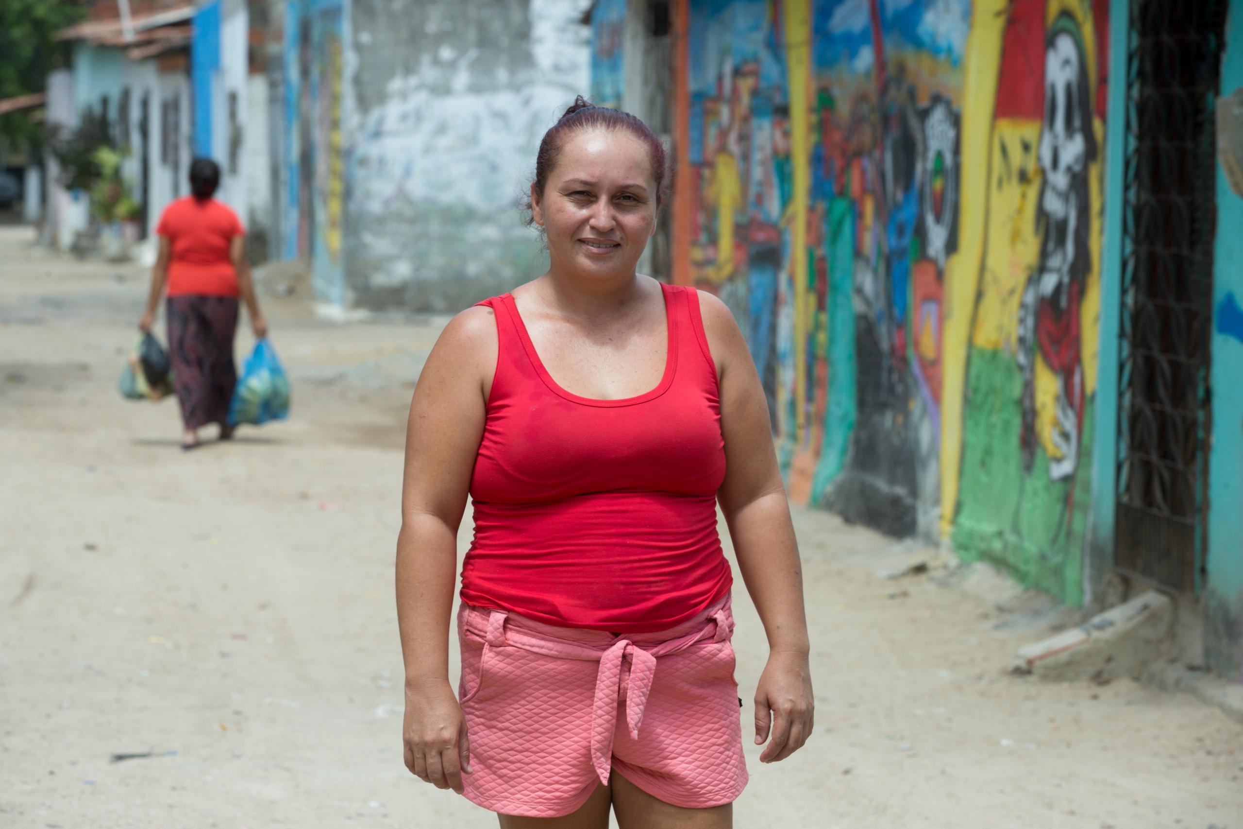 Retrato de uma mulher em pé em uma rua de comunidade, com paredes coloridas de grafite e outra pessoa carregando sacolas ao longe.
