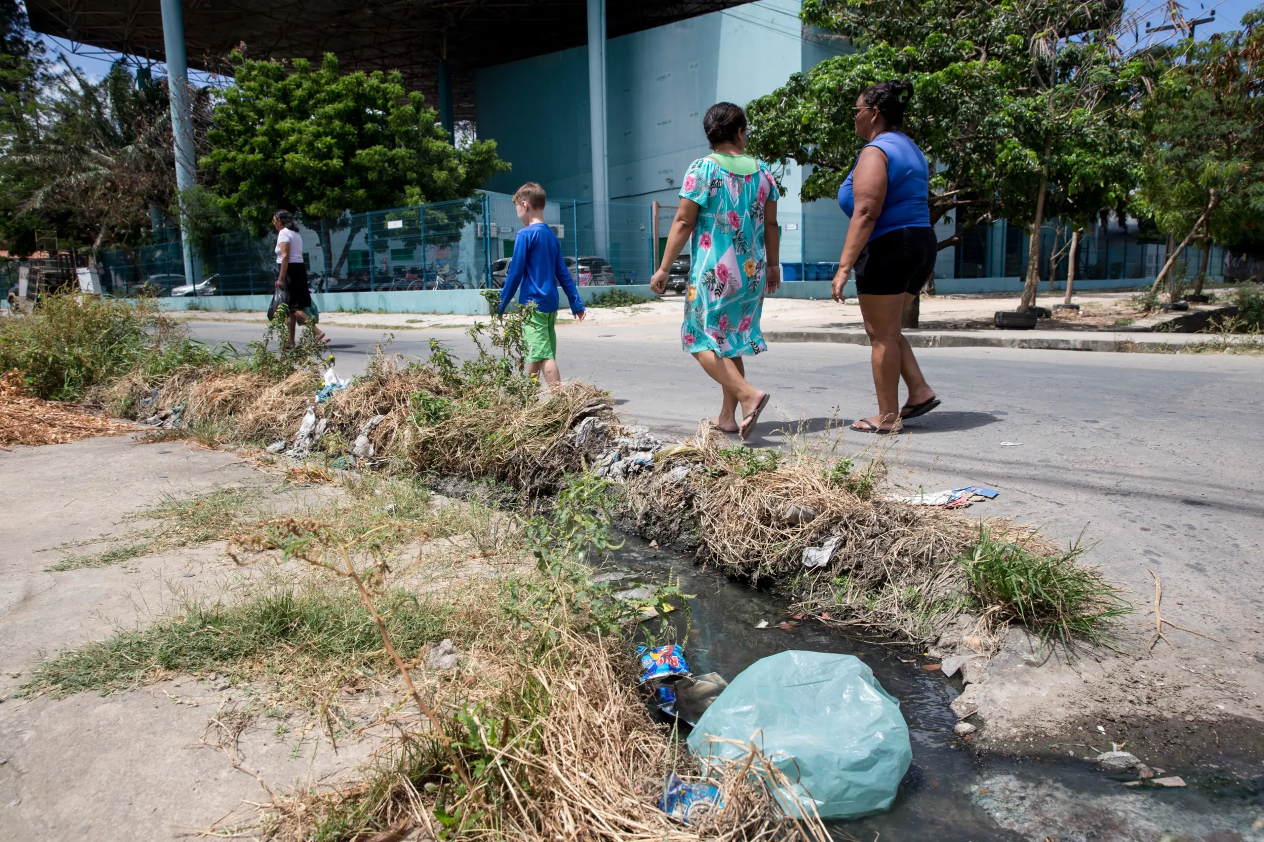Quatro pessoas caminham em uma calçada ao lado de uma sarjeta com lixo, mato seco, esgoto e entulho, com um saco plástico azul claro no primeiro plano.