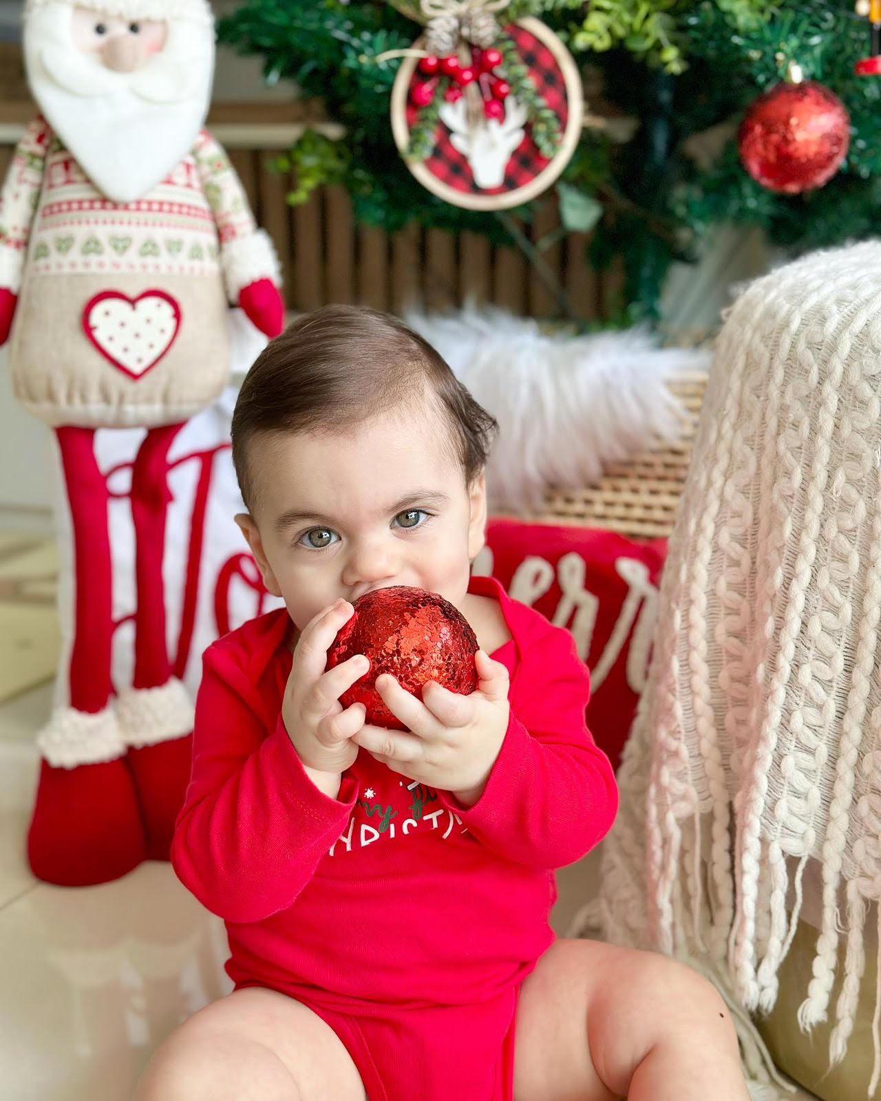 Um bebê de olhos verdes/azulados e cabelo castanho, vestindo um body vermelho com detalhes brancos de Natal, sentado no chão e segurando uma grande bola de Natal vermelha e brilhante perto da boca. O bebê olha diretamente para a câmera com uma expressão curiosa. Ao fundo, um ambiente decorado para o Natal, com um Papai Noel de pano grande, uma almofada vermelha com a palavra 