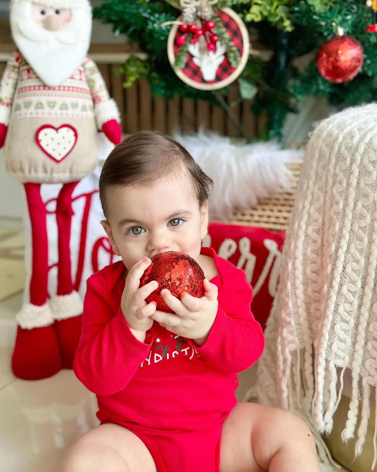Um bebê de olhos verdes/azulados e cabelo castanho, vestindo um body vermelho com detalhes brancos de Natal, sentado no chão e segurando uma grande bola de Natal vermelha e brilhante perto da boca. O bebê olha diretamente para a câmera com uma expressão curiosa. Ao fundo, um ambiente decorado para o Natal, com um Papai Noel de pano grande, uma almofada vermelha com a palavra 