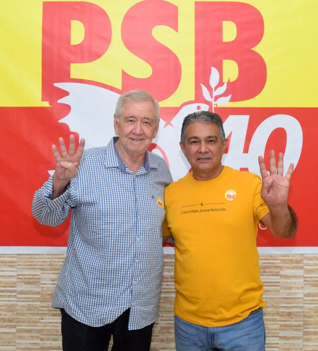 Moésio Loiola e Solano Feitosa posam lado a lado diante de um banner do PSB. Ambos sorriem e levantam uma das mãos, mostrando quatro dedos. Um usa camisa xadrez azul; o outro veste camiseta amarela com adesivo do partido.