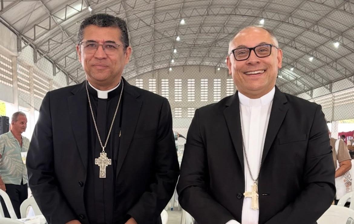 Dois bispos em traje clerical, Dom Antônio Carlos (esquerda) e Dom Gregório (direita), posam lado a lado e sorrindo, usando cruzes peitorais grandes e prateadas. Eles estão em um ambiente amplo, possivelmente uma área de eventos coberta, com pessoas em segundo plano.