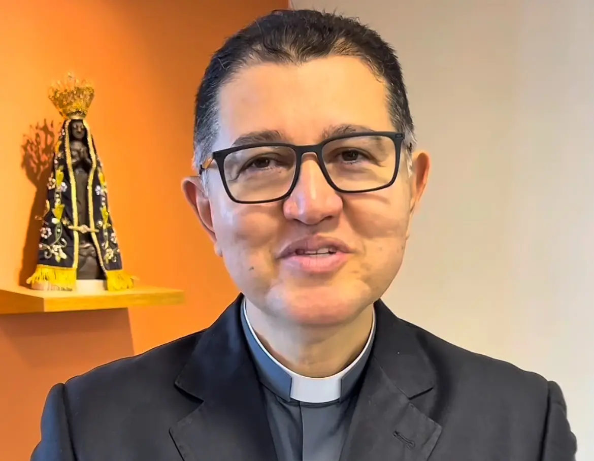 Um retrato em close-up de Dom Jânison de Sá, que sorri levemente e usa óculos e traje clerical (camisa preta com colarinho branco). Ele está em um fundo laranja/amarelo, e sobre uma prateleira à sua esquerda está uma imagem de Nossa Senhora Aparecida.