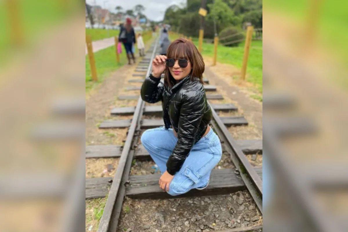 Um retrato de corpo inteiro de uma jovem identificada como Taynara Souza, agachada sobre trilhos de trem em uma área externa. Ela usa calça jeans clara, uma jaqueta preta de nylon e óculos de sol, olhando para a câmera e sorrindo. O fundo, ligeiramente desfocado, mostra mais pessoas e vegetação.