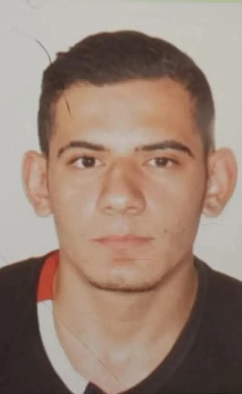 Uma foto de identificação (tipo 3x4 ou documento) de um homem jovem, Douglas Alves da Silva. Ele tem cabelo escuro curto e orelhas proeminentes, olhando diretamente para a câmera com uma expressão neutra. Ele veste uma camiseta escura com gola em V e detalhes claros.