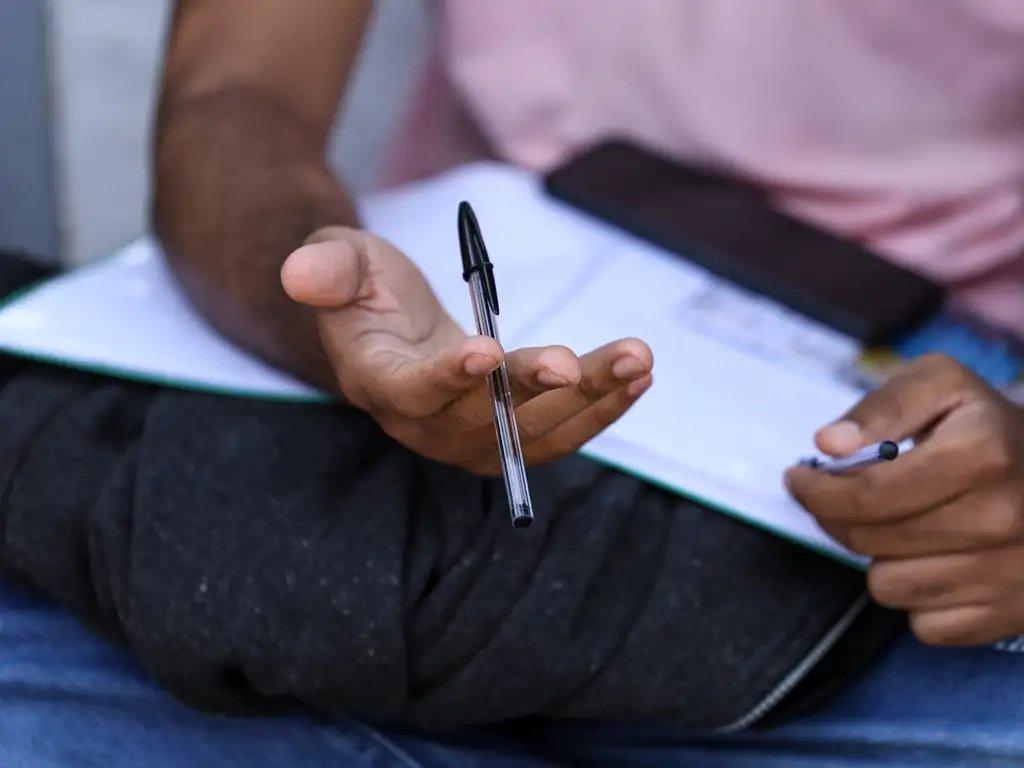 Mão de pessoa negra segurando caneta esferográfica de tampa preta. Ao fundo, borrados em sua perna, papel branco de prova e celular preto.