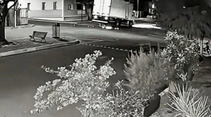 Câmera de segurança noturna em preto e branco mostrando um caminhão com as rodas esquerdas suspensas em uma rua urbana com banco e arbustos em primeiro plano.