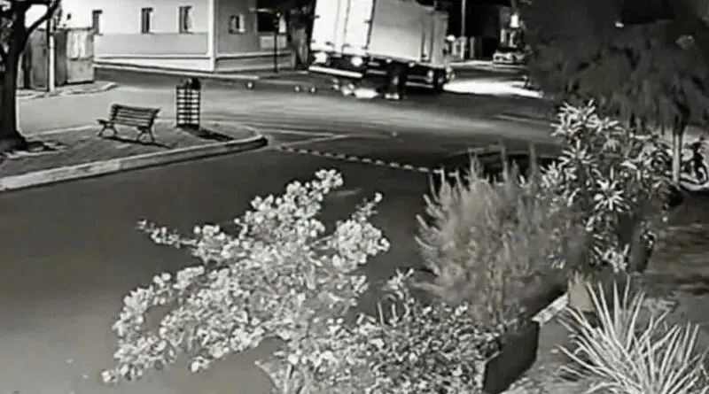 Câmera de segurança noturna em preto e branco mostrando um caminhão com as rodas esquerdas suspensas em uma rua urbana com banco e arbustos em primeiro plano.