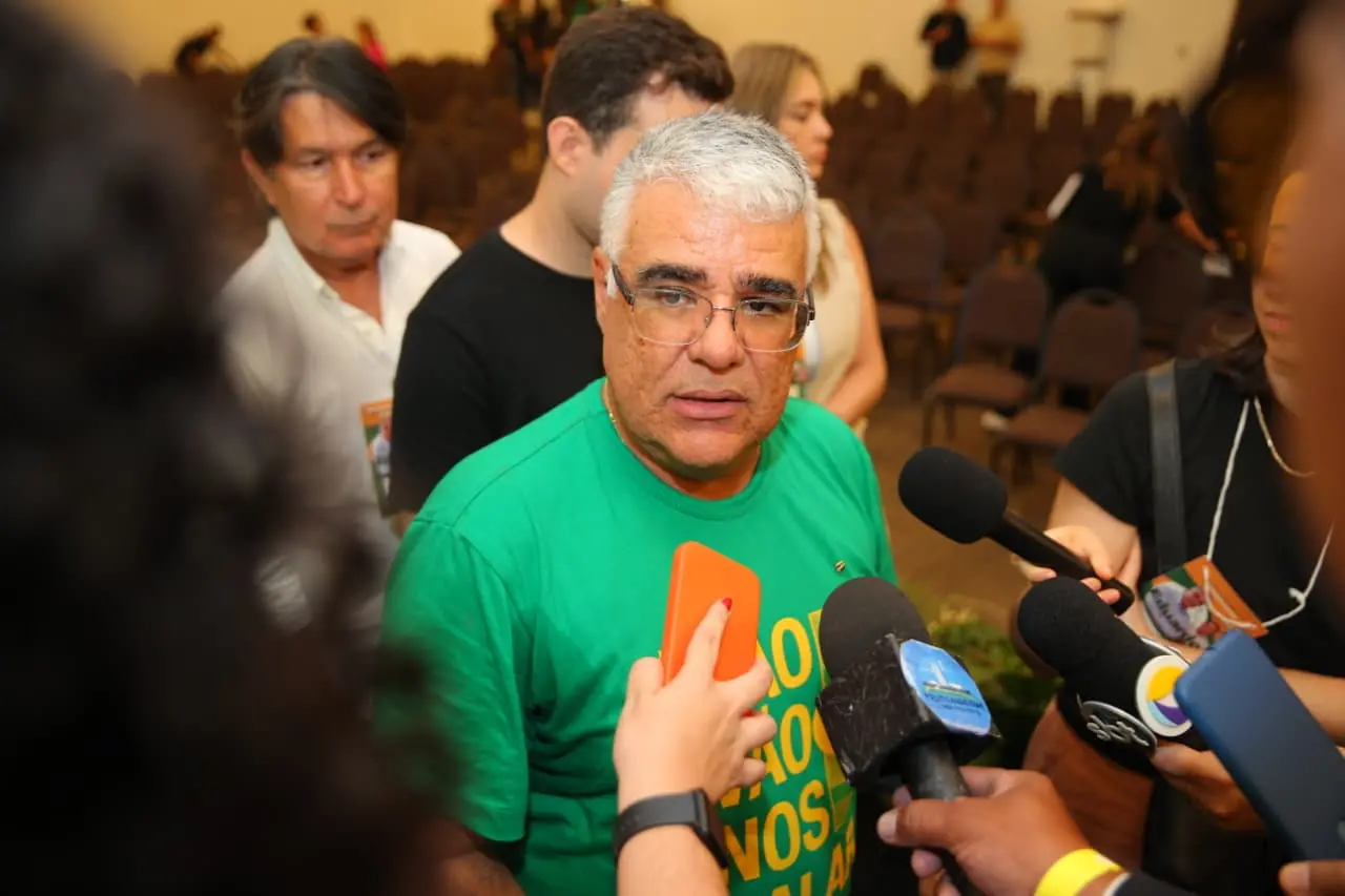 Eduardo Girão de camiseta verde fala com a imprensa em um auditório. Ele está cercado por vários microfones e jornalistas, enquanto outras pessoas aparecem desfocadas ao fundo.