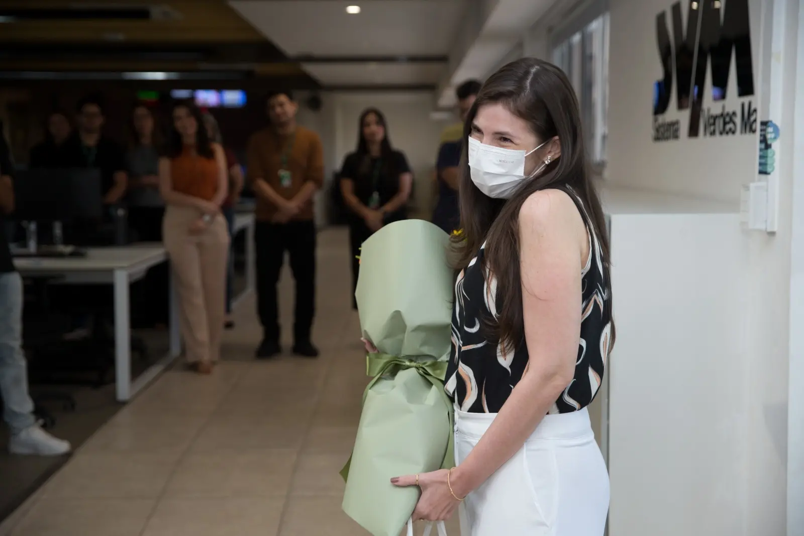 Marina Alves com máscara sorri e segura um buquê de flores embrulhado em verde, em um ambiente de escritório com colegas de trabalho desfocados ao fundo e o logo Sistema Verdes Mares.