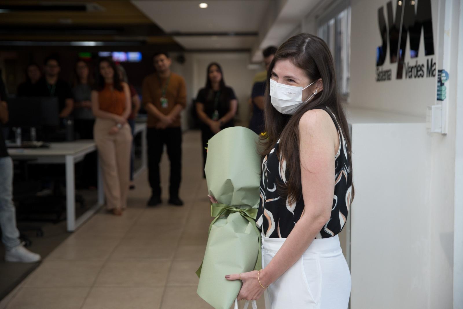 Marina Alves com máscara sorri e segura um buquê de flores embrulhado em verde, em um ambiente de escritório com colegas de trabalho desfocados ao fundo e o logo Sistema Verdes Mares.