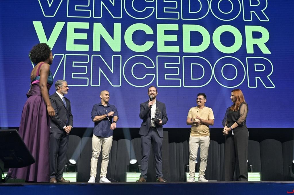 A imagem registra um grupo de seis pessoas no palco durante uma premiação. Ao fundo, um grande painel azul exibe repetidamente a palavra “VENCEDOR” em letras grandes. No centro, um homem de terno segura um troféu e fala ao microfone.
