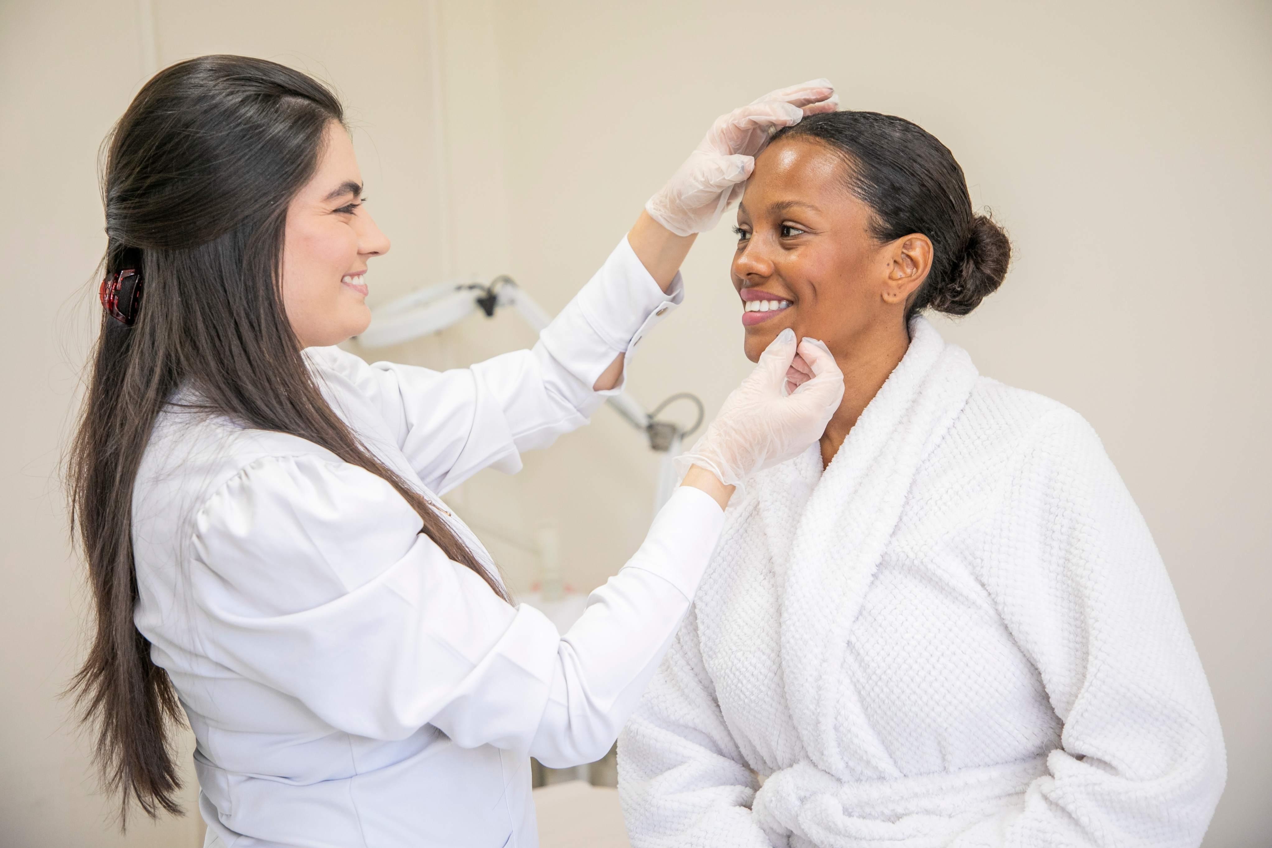 Foto de uma profissional da área da saúde ou estética, vestindo jaleco branco e luvas descartáveis, realiza uma avaliação facial em uma mulher que está sentada, usando um roupão branco. A profissional sorri enquanto toca delicadamente a testa e o queixo da paciente, que também sorri de forma tranquila. O ambiente é claro, com aparência de consultório ou clínica estética.