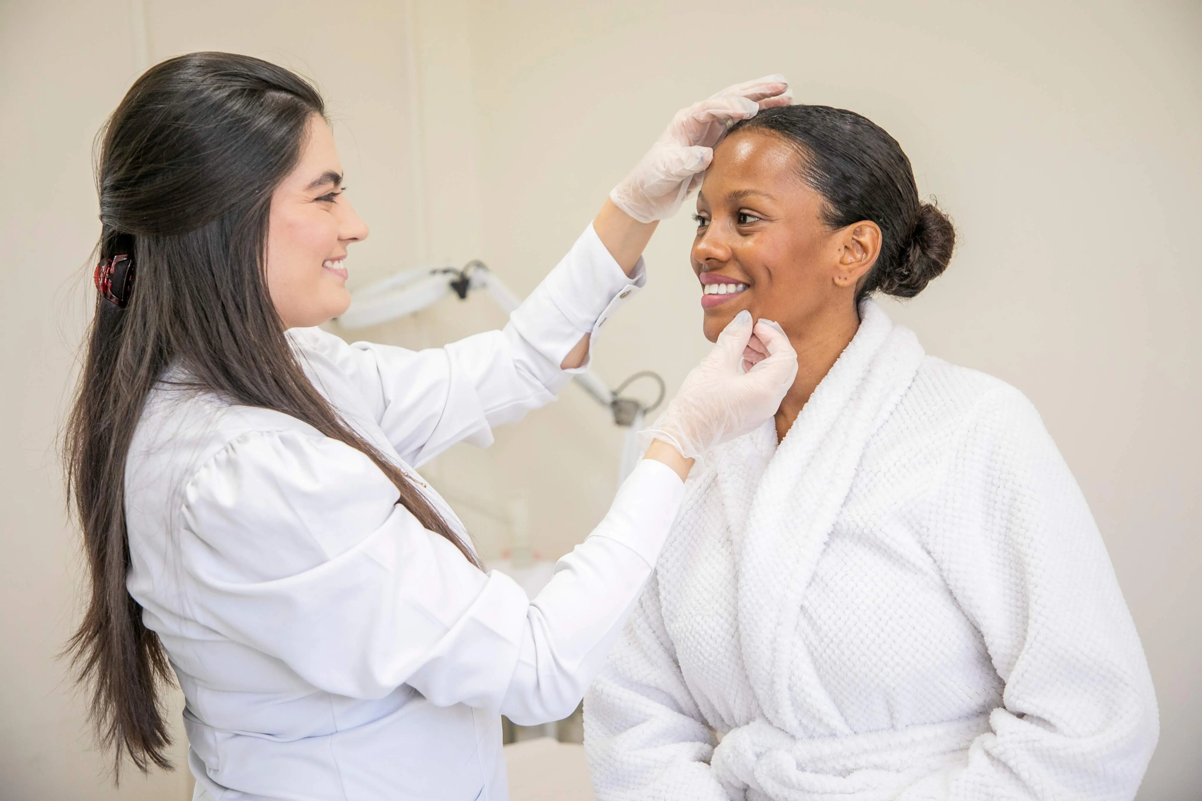 Foto de uma profissional da área da saúde ou estética, vestindo jaleco branco e luvas descartáveis, realiza uma avaliação facial em uma mulher que está sentada, usando um roupão branco. A profissional sorri enquanto toca delicadamente a testa e o queixo da paciente, que também sorri de forma tranquila. O ambiente é claro, com aparência de consultório ou clínica estética.