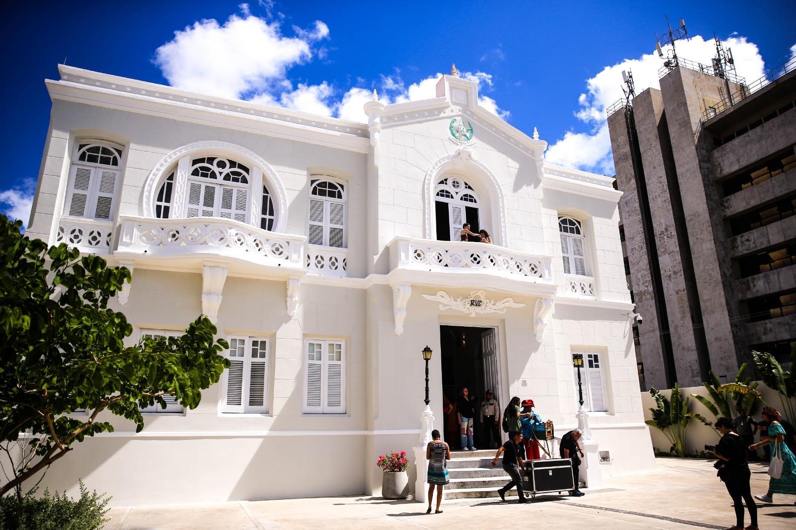Na imagem, uma fotografia externa de um edifício branco imponente de dois andares com arquitetura clássica, possivelmente uma construção histórica reformada. Apresenta uma varanda principal (sacada) com grades de balaústres brancos rendilhados e duas portas-janelas em arco, ambas com venezianas brancas fechadas. Uma segunda varanda menor, central, acima da entrada principal, também possui balaústres semelhantes, e uma pessoa está olhando para fora.