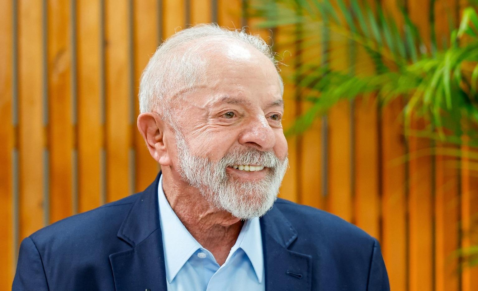 Lula de perfil. Ele é um homem branco de cabelo branco e barba da mesma cor. Na foto, ele está sorrindo e usa terno azul e camisa social azul clara.