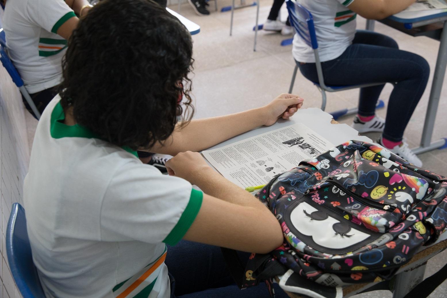 Jovem sentada em carteira escolar com uma mochila colorida ao lado, focado na leitura de material impresso durante a aula.