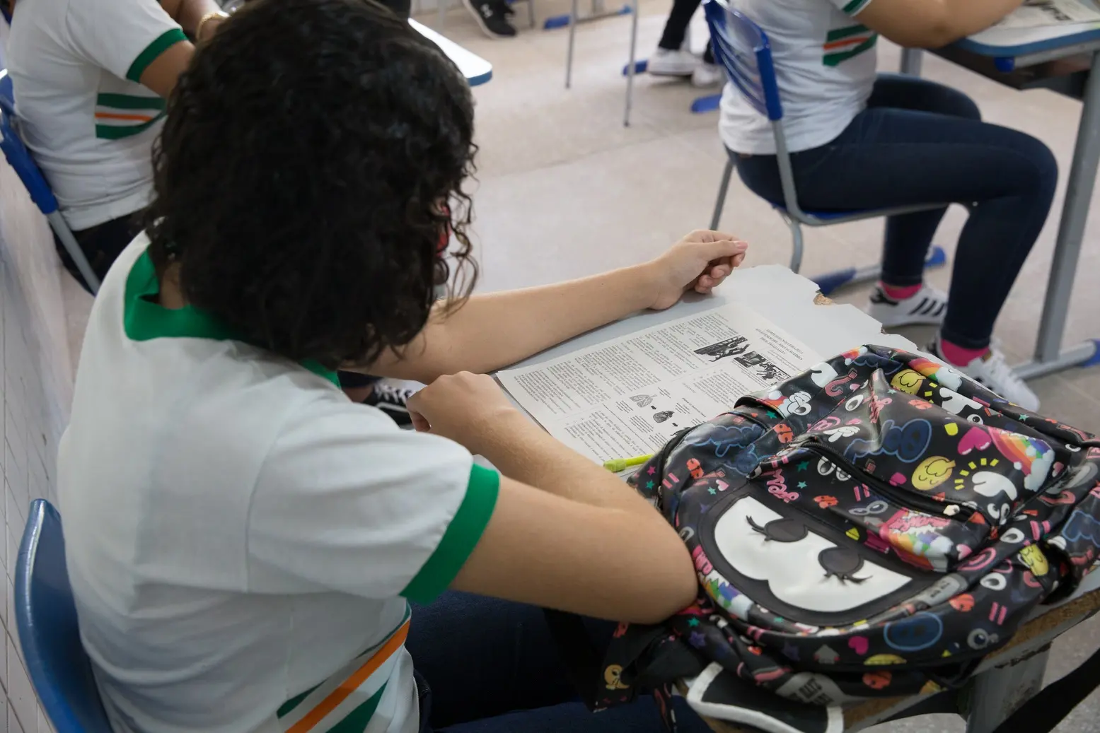 Jovem sentada em carteira escolar com uma mochila colorida ao lado, focado na leitura de material impresso durante a aula.