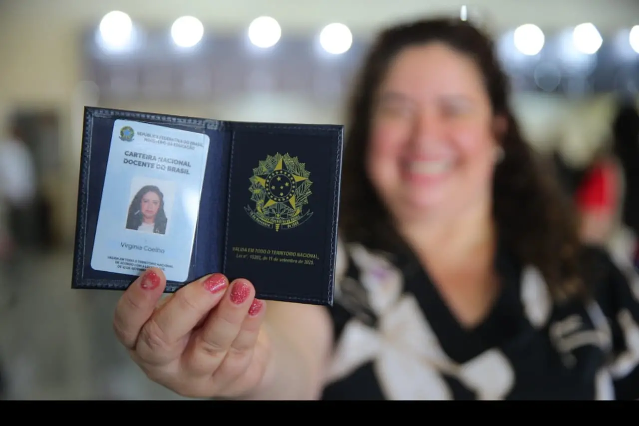 Mão de uma mulher segurando a Carteira Nacional Docente do Brasil, com foto e o brasão da República, enquanto uma mulher sorri desfocada ao fundo.