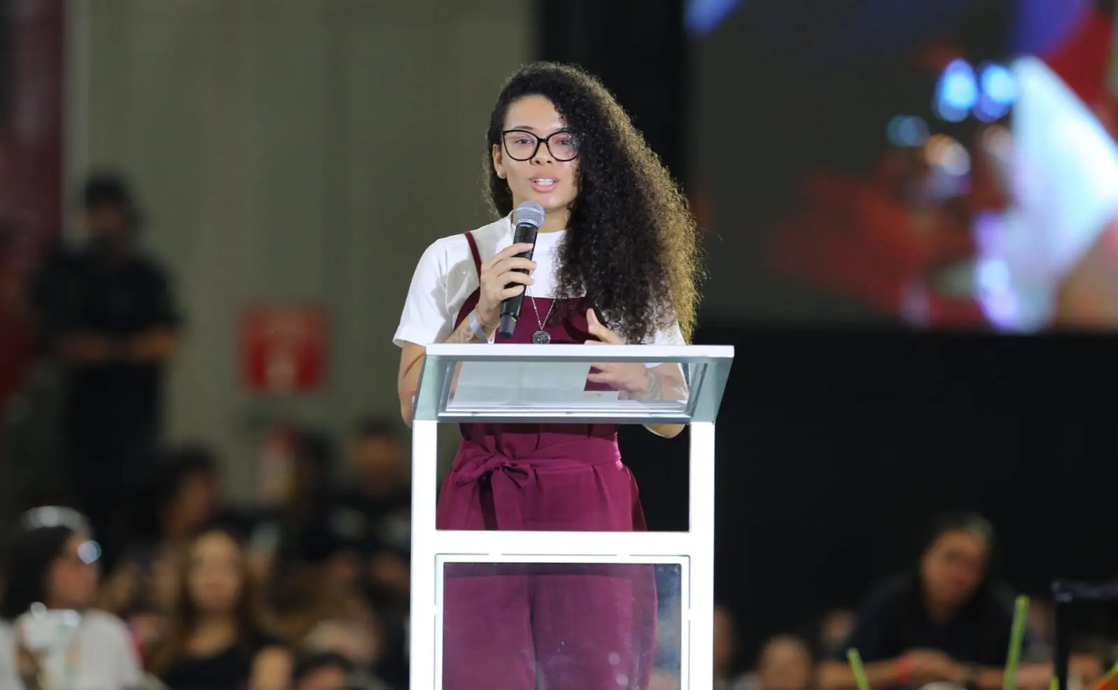 Jovem negra de cabelos longos cacheados, vestindo roupa vinho e branca, discursa em púlpito no Centro de Eventos do Ceará.
