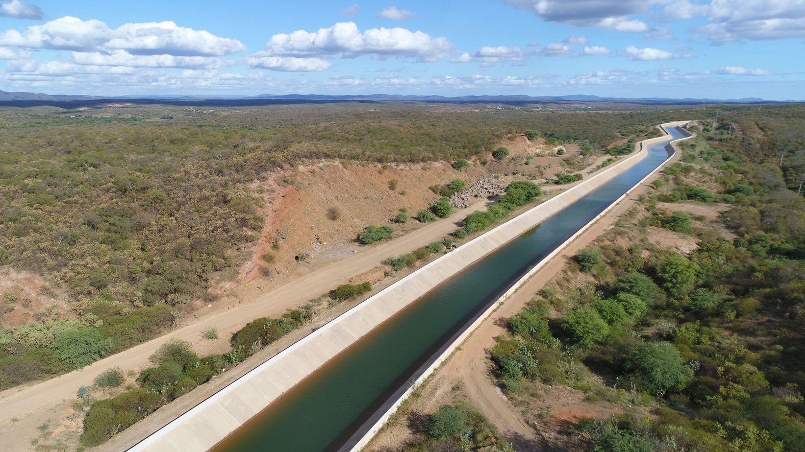 Vista aérea de um longo canal de concreto com água verde-azulada, atravessando uma paisagem semiárida com vegetação esparsa e estradas de terra sob um céu azul com nuvens. O canal é da Transposição do Rio São Francisco no Ceará.