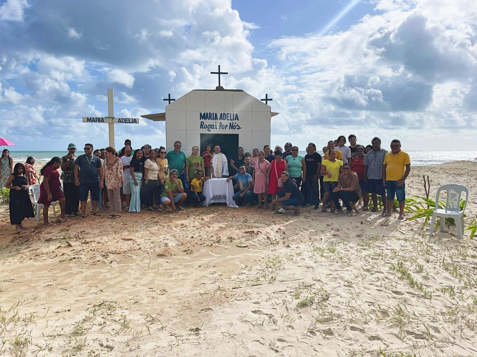 A foto mostra um grande grupo de moradores reunidos em frente à capelinha dedicada a Maria Adélia, construída na faixa de areia entre Mundaú e Flecheiras. Ao centro, um celebrante está diante de uma pequena mesa coberta por um pano branco, indicada para a missa realizada no local.