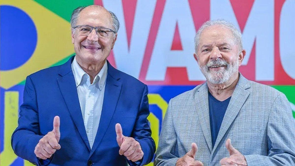 Duas pessoas idosas posam lado a lado sorrindo, ambas fazendo o gesto de positivo com as mãos. A pessoa à esquerda veste um terno azul e óculos; a pessoa à direita usa um blazer cinza claro sobre uma camisa escura. Ao fundo, há um painel colorido com elementos da bandeira do Brasil e grandes letras vermelhas.