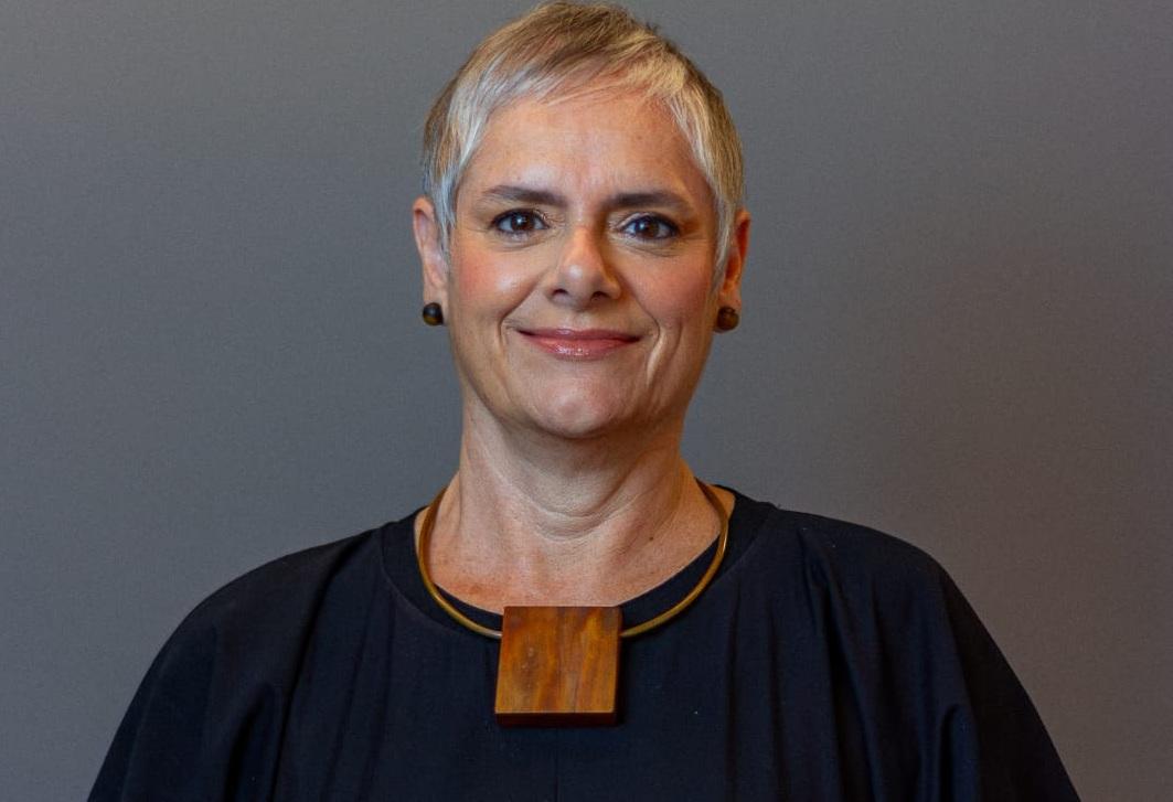 A imagem mostra uma mulher adulta, de cabelos curtos e grisalhos, usando brincos pequenos e um colar com um pingente de madeira. Ela veste uma blusa escura e sorri levemente, olhando diretamente para a câmera. O fundo é neutro, em tom acinzentado, destacando sua expressão serena e profissional.