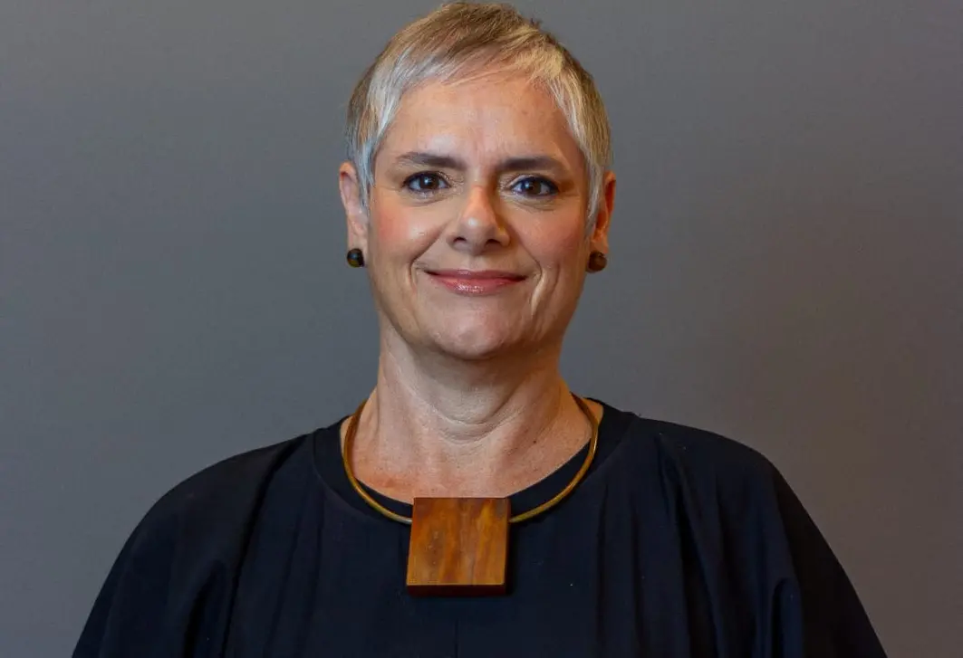 A imagem mostra uma mulher adulta, de cabelos curtos e grisalhos, usando brincos pequenos e um colar com um pingente de madeira. Ela veste uma blusa escura e sorri levemente, olhando diretamente para a câmera. O fundo é neutro, em tom acinzentado, destacando sua expressão serena e profissional.