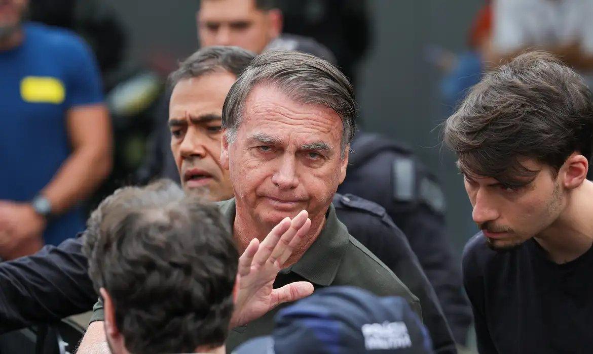 Bolsonaro cercado por agentes de segurança, com uniformes e coletes táticos visíveis, em ambiente externo.