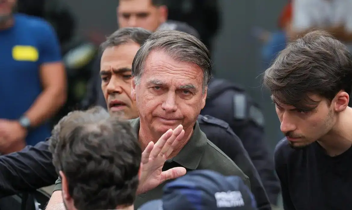 Bolsonaro cercado por agentes de segurança, com uniformes e coletes táticos visíveis, em ambiente externo.