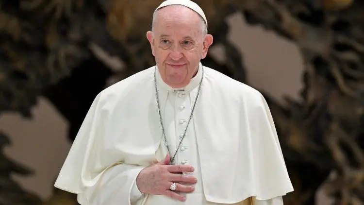 Papa Francisco em traje papal branco, com um sorriso gentil e olhar atento, durante uma audiência no Vaticano. Ele usa um crucifixo pendurado e um anel no dedo anelar direito.