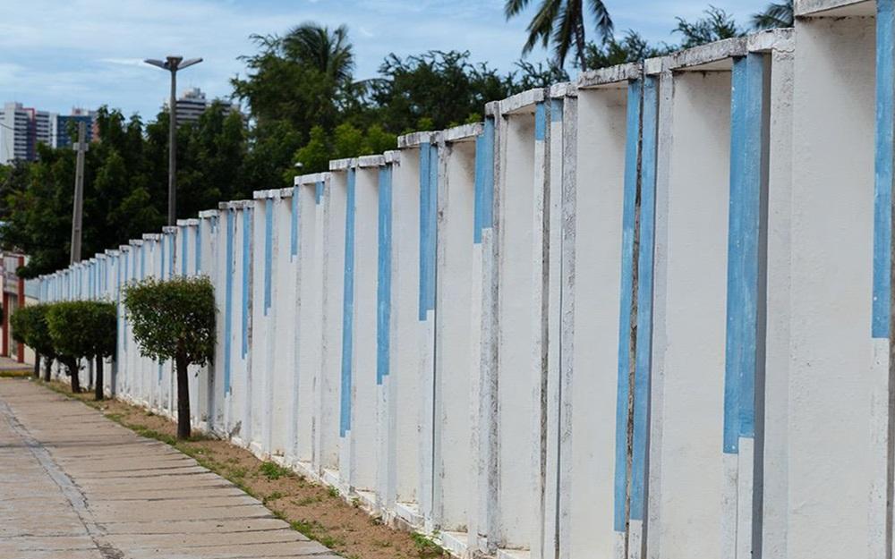 Na imagem, uma longa parede branca de concreto, com uma série de pilares verticais e reentrâncias. Dentro das reentrâncias, faixas verticais pintadas de azul-claro podem ser vistas, criando um efeito de arte cinética ou ilusão de ótica quando o observador se move. A parede se estende pela imagem, ladeando um calçadão à esquerda onde pequenos arbustos quadrados estão plantados. No fundo, há árvores altas, palmeiras e edifícios urbanos, sob um céu nublado. A imagem é creditada a Gentil Barreira.