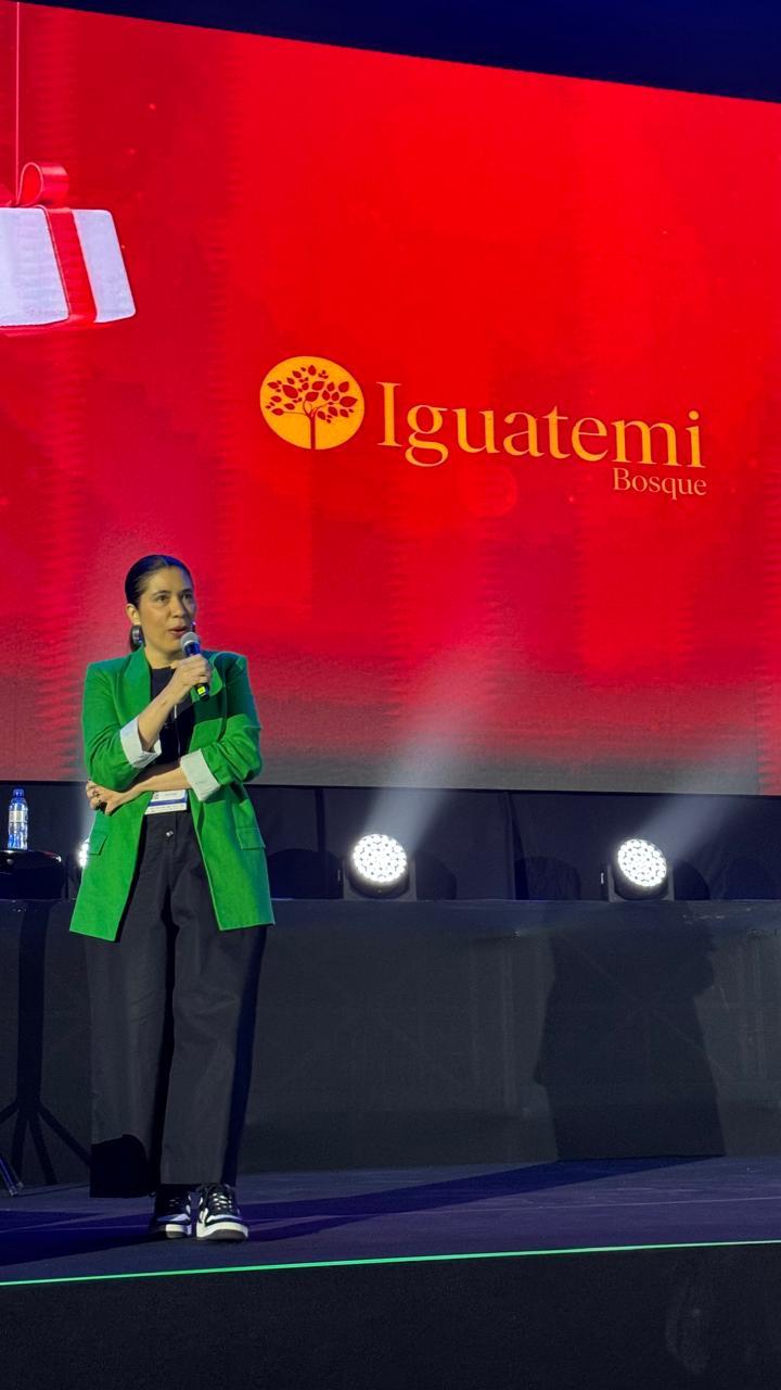 A imagem mostra uma mulher em um palco, usando blazer verde e segurando um microfone. Atrás dela, um grande painel vermelho exibe o nome e o logo de um shopping. O cenário sugere uma apresentação ou palestra.
