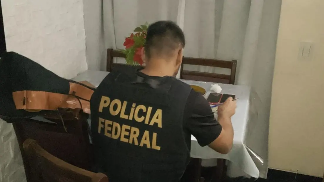 A imagem mostra um policial federal de costas, fardado e sentado em uma mesa, enquanto realiza uma busca em uma operação policial.