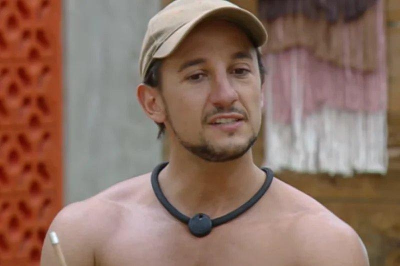 Pessoa usando boné claro e colar preto com dispositivo circular, em ambiente rústico com parede decorativa vermelha e painel têxtil ao fundo.