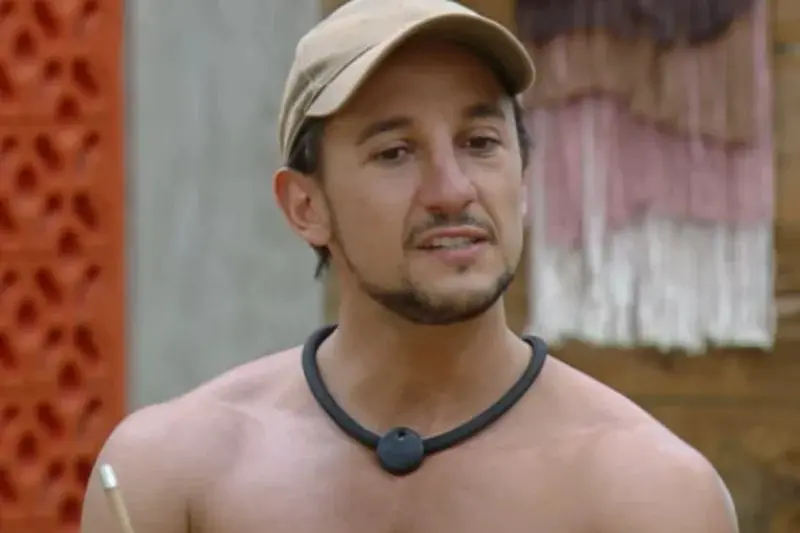 Pessoa usando boné claro e colar preto com dispositivo circular, em ambiente rústico com parede decorativa vermelha e painel têxtil ao fundo.