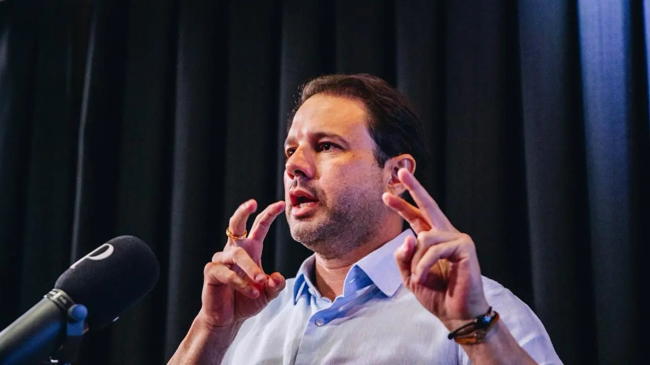 Leo Couto faz gesto de aspas com as mãos. Ele está sentado, diante dele há um microfone e, ao fundo, um tecido preto.